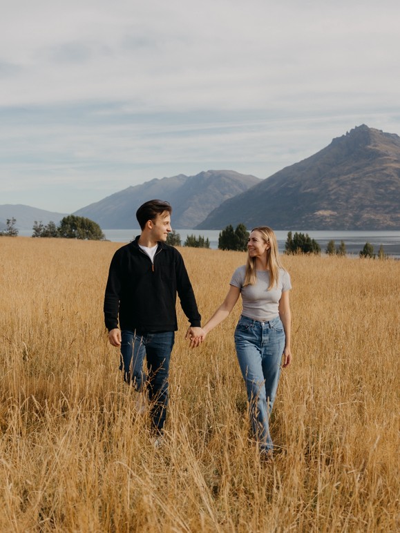 Matia and Jillian walking hand in hand through tall golden grass, with mountains and a lake behind them